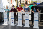 Pięć młodych osób stoi przy białych mównicach i trzyma w rękach chorągiewki z flagami Unii Europejskiej.