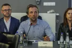 Mężczyzna siedzący przy stole konferencyjnym przemawia do mikrofonu