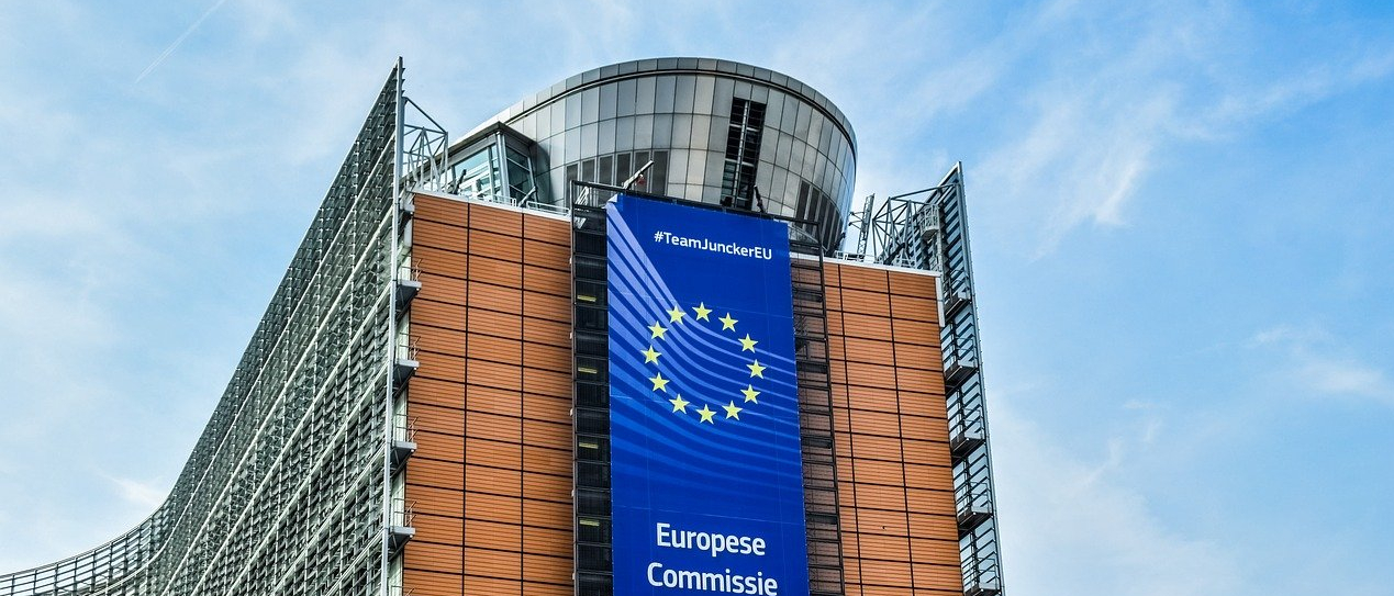 Budynek Komisji Europejskiej. Architektura budynku współczesna. Na środku zdjęcia pomarańczowa ściana z flagą UE i napisem Komisja Europejska. Po bokach, fasada ze szkła i stali w kształcie łuku.