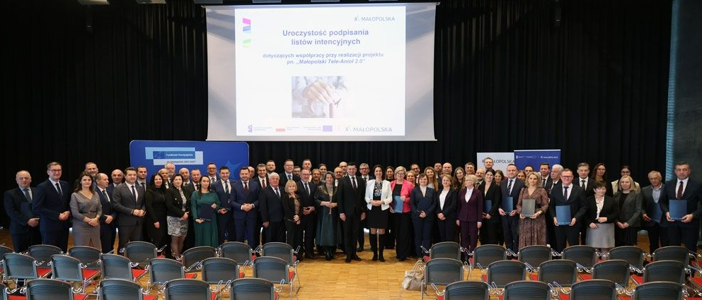 W centrum kadru grupa ludzi pozująca do zdjęcia. Nad nimi duży ekran z wyświetloną prezentacją. U dołu kadru kilka rzędów krzeseł.