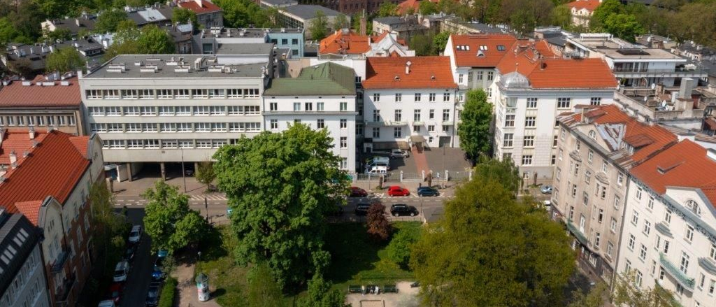 Budynek szpitalny w otoczeniu kamienic i zieleni.