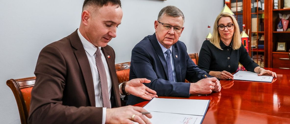 Mężczyzna podpisujący umowę. Za nim wicemarszałek Witold Kozłowski i kobieta w blond włosach.