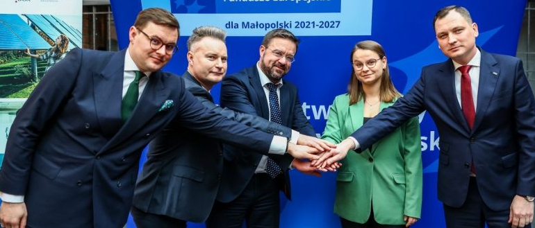 Pięć osób w formalnych strojach stoi przed niebieskim tłem z napisem „Fundusze Europejskie dla Małopolski 2021-2027”. Wyciągają ręce w centralnym stosie, pokazując gest współpracy.