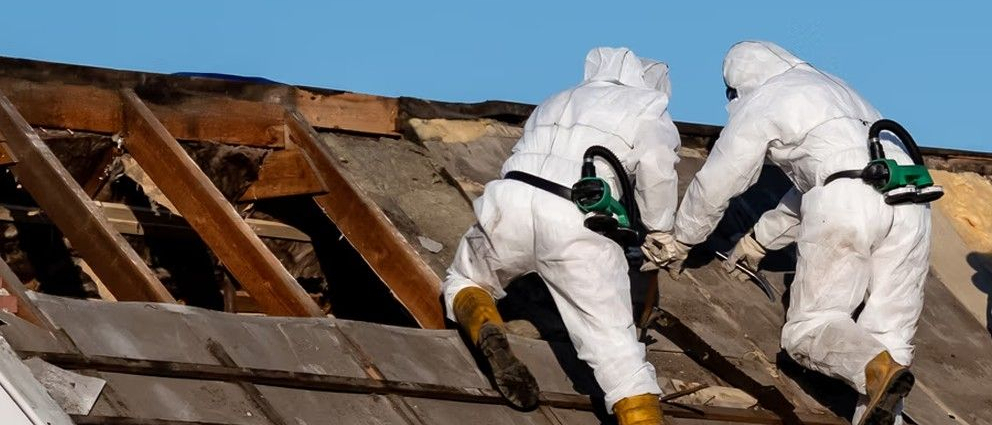 Robotnicy w kombinezonach ochronnych ściągają azbest z dachu.