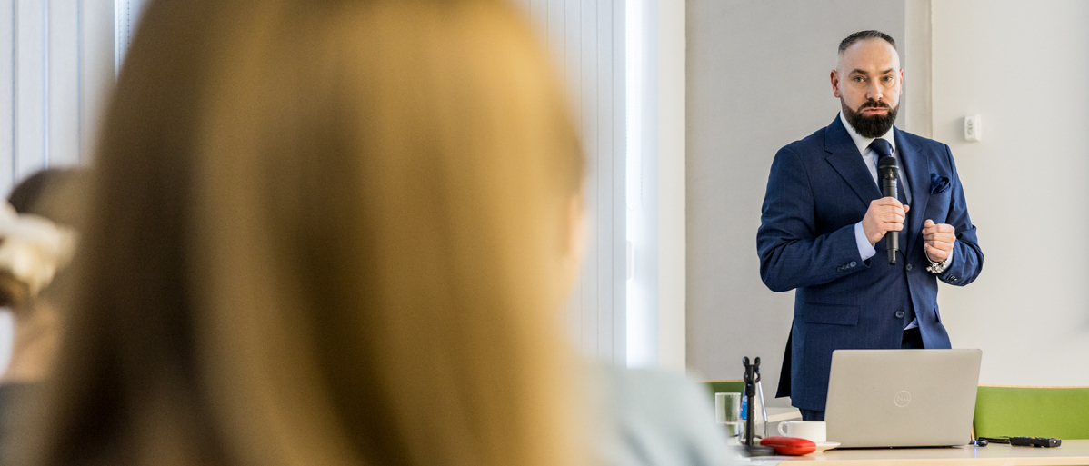 Na pierwszym planie ujęcie od tyłu grupę osób siedzących na krzesłach w sali szkolenowej. W głębi kadru prelegent trzymający w dłoni mikrofon