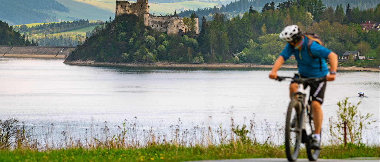 Mężczyzna jadący na rowerze. W tle jezioro, zamek i góry