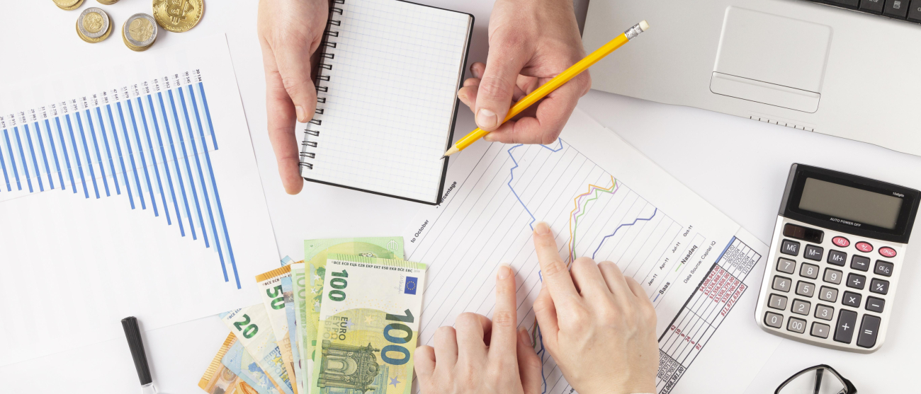 Widoczne dłonie dwóch osób na biurku. Rozłożone dokumenty, wykresy, monety, banknoty euro, kalkulator, okulary, fragment tabletu.