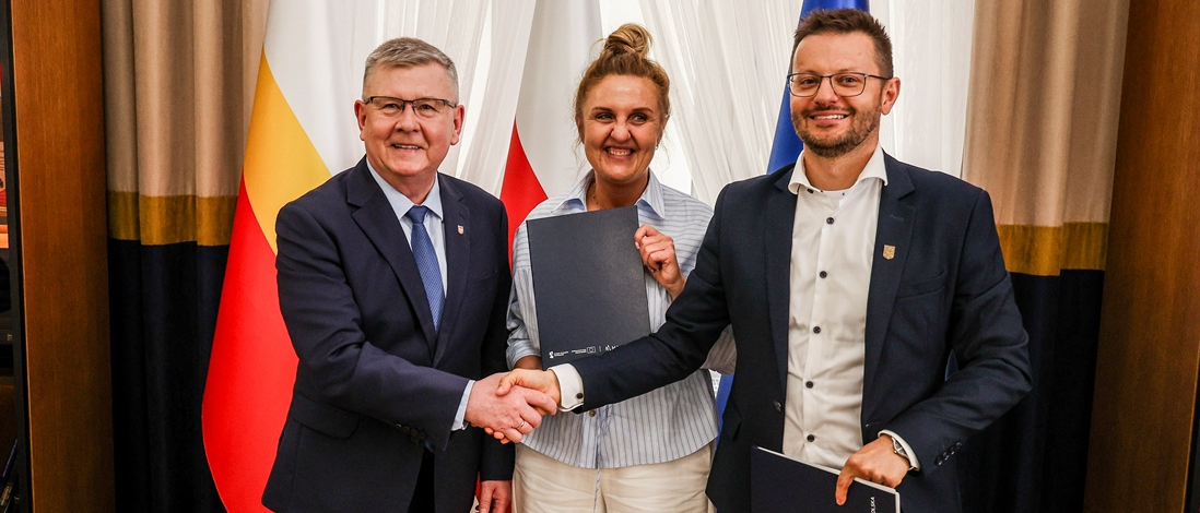 Witold Kozłowski i Bartosz Kaliński ściskają swoje dłownie tuż po podpisaniu umowy. Kinga Gurdek trzyma okolicznościową teczkę z dokumentami. Cała trójka stoi na tle flag Małopolski, Polski i Unii Europejskich